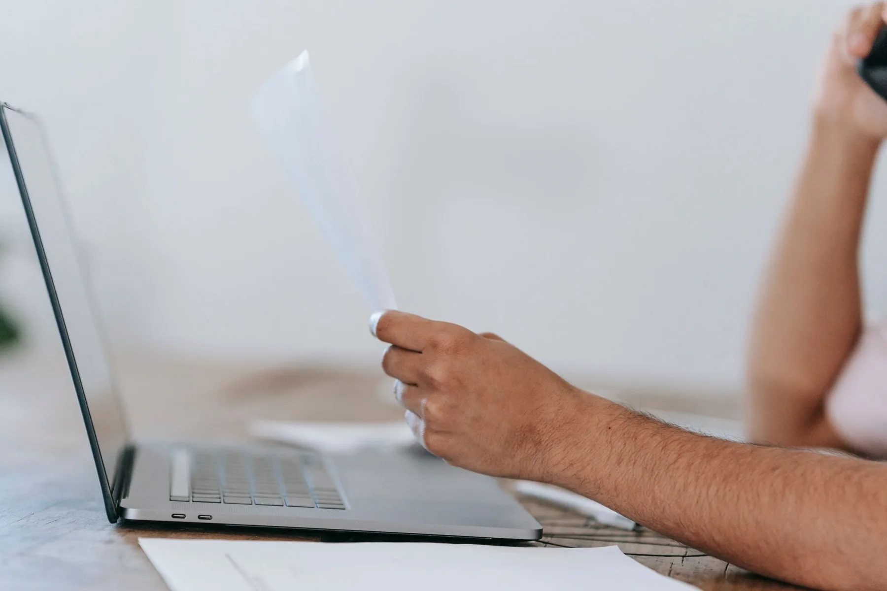 A person reviews paperwork beside an open laptop while holding a phone.