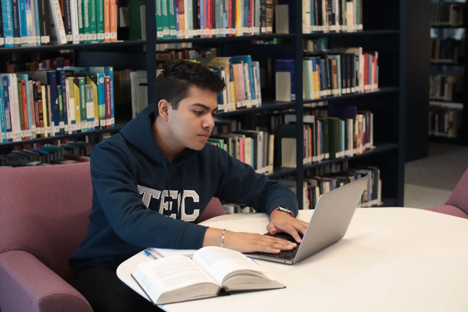 A student studies at a library table with a laptop, open book, and notes.
