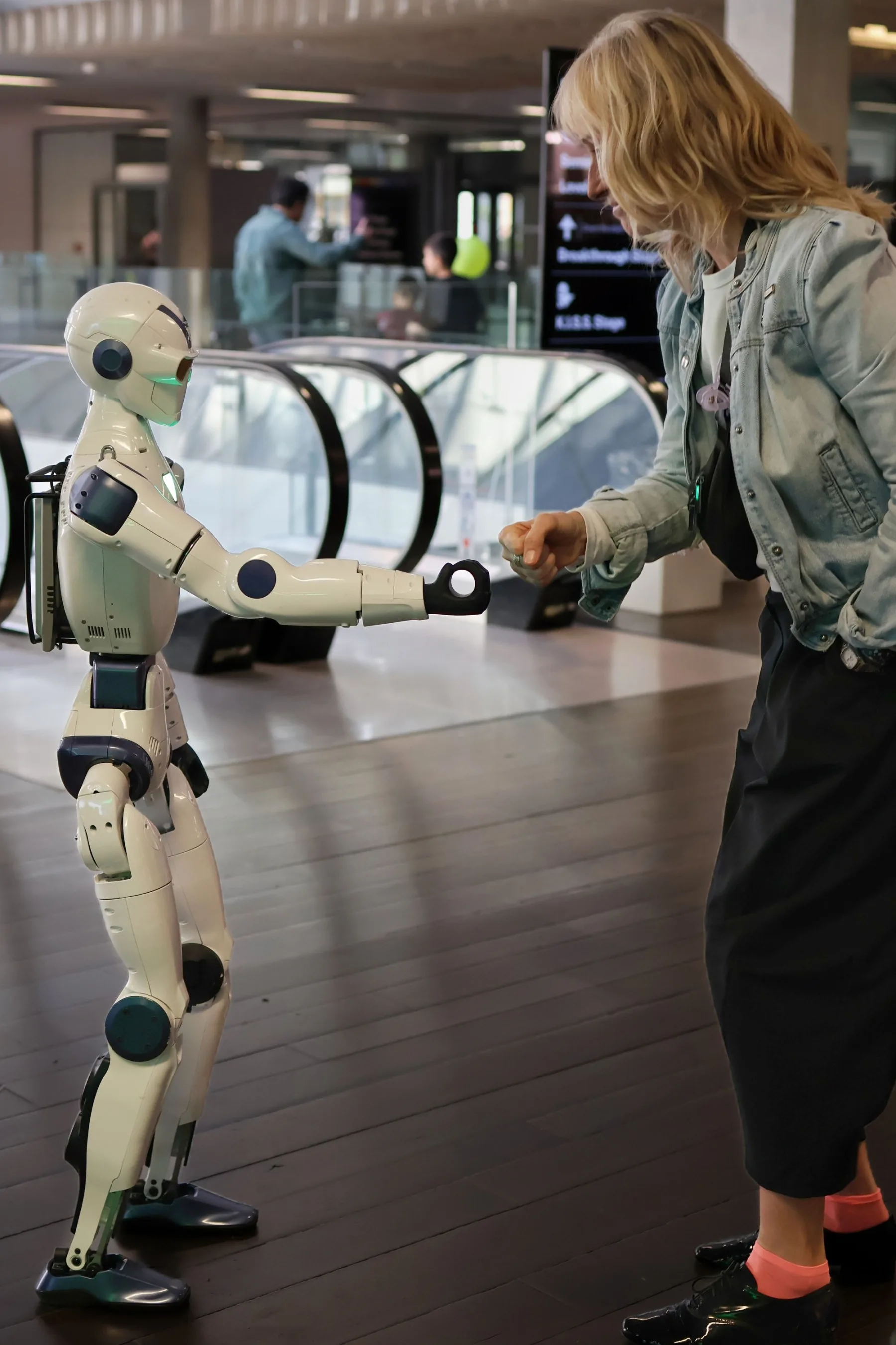 A humanoid robot extends a hand toward a person in a public indoor setting.